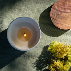 White ceramic candle vessel with glowing flame casting shadows, accompanied by green moss and wooden candle lid on textured surface