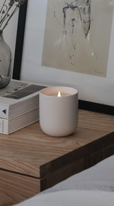 White ceramic candle burning on wooden nightstand next to books and vase in cozy bedroom setting