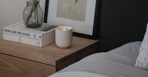 White ceramic candle burning on wooden nightstand next to books and vase in cozy bedroom setting