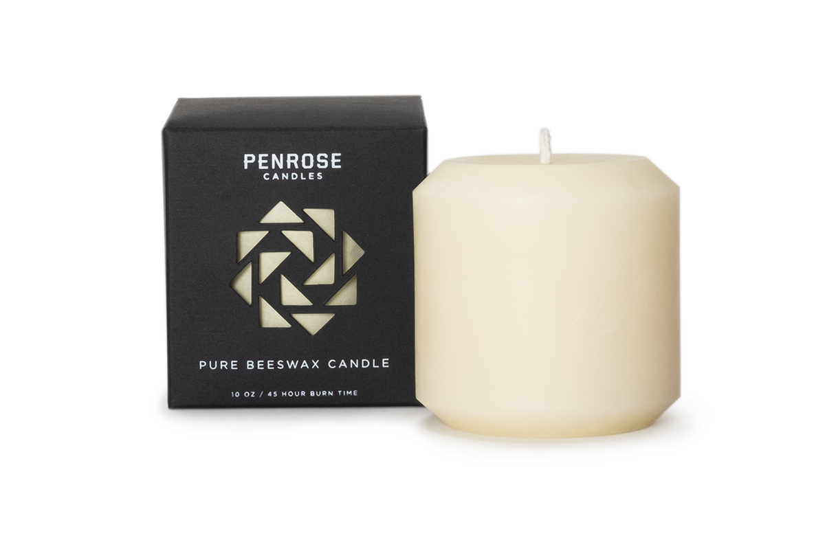 A natural 3 inch  ivory pillar candle next to its black packaging box with a geometric pattern, labeled as 'Penrose Pure Beeswax Candle'.